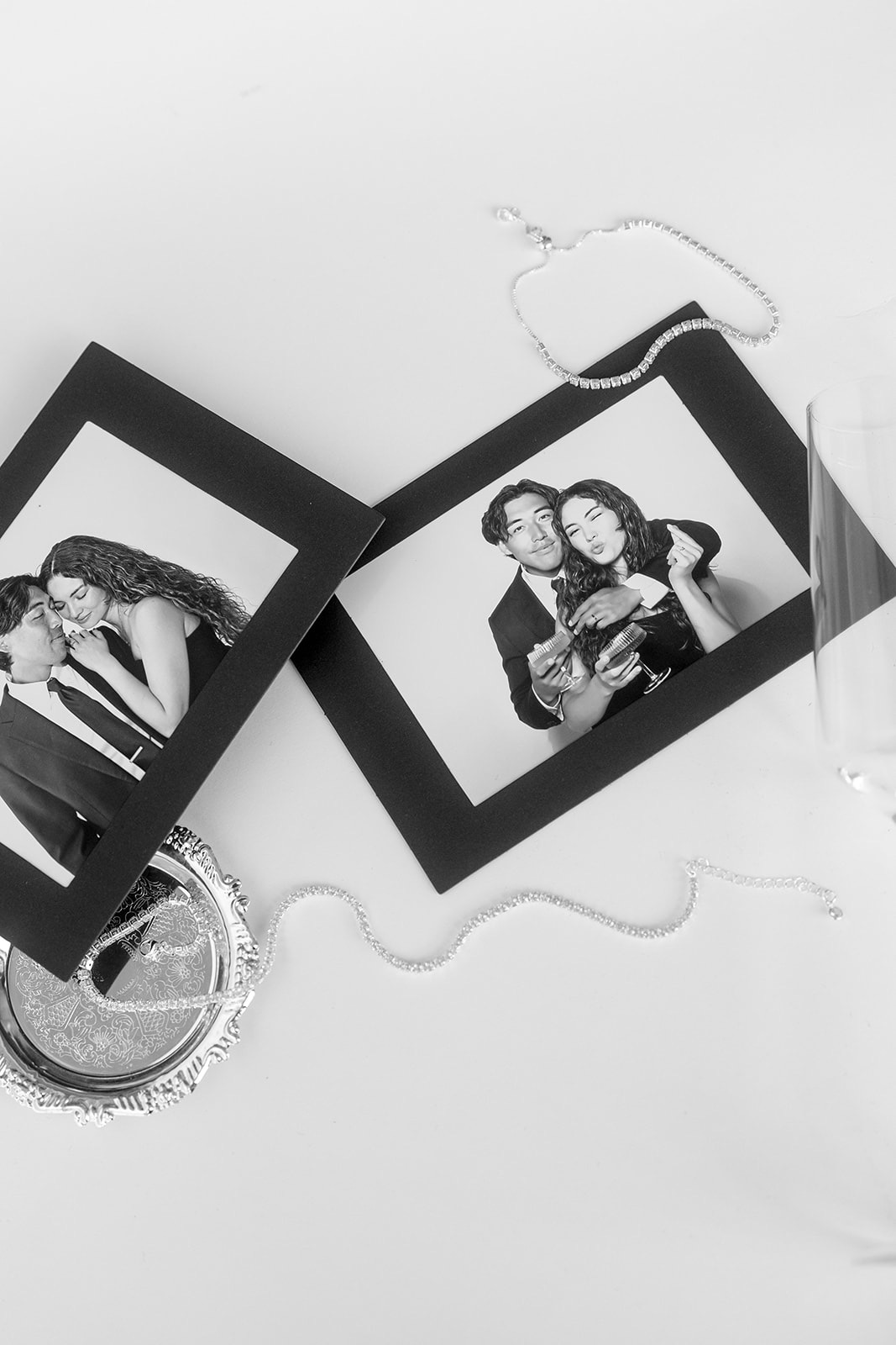 Black-and-white flat lay of two framed photo booth prints of a couple beside an ornate tray and sparkly jewelry