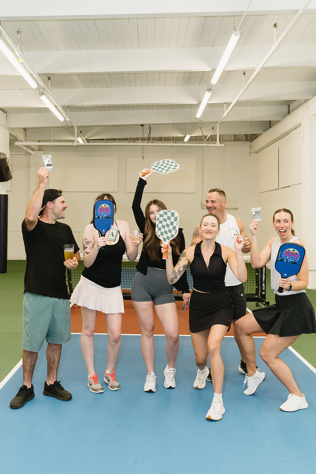 Six people on an indoor pickleball court raise paddles and small photo prints