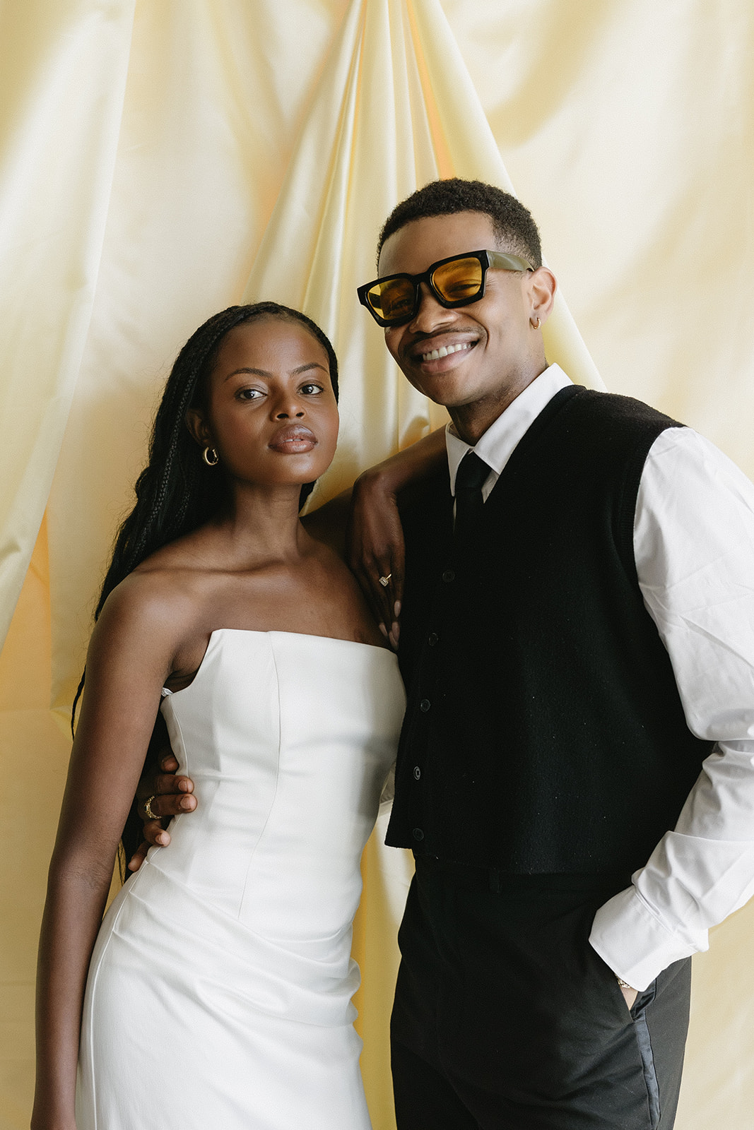 A couple at a cream draped backdrop, she in a strapless white dress, he in tinted sunglasses