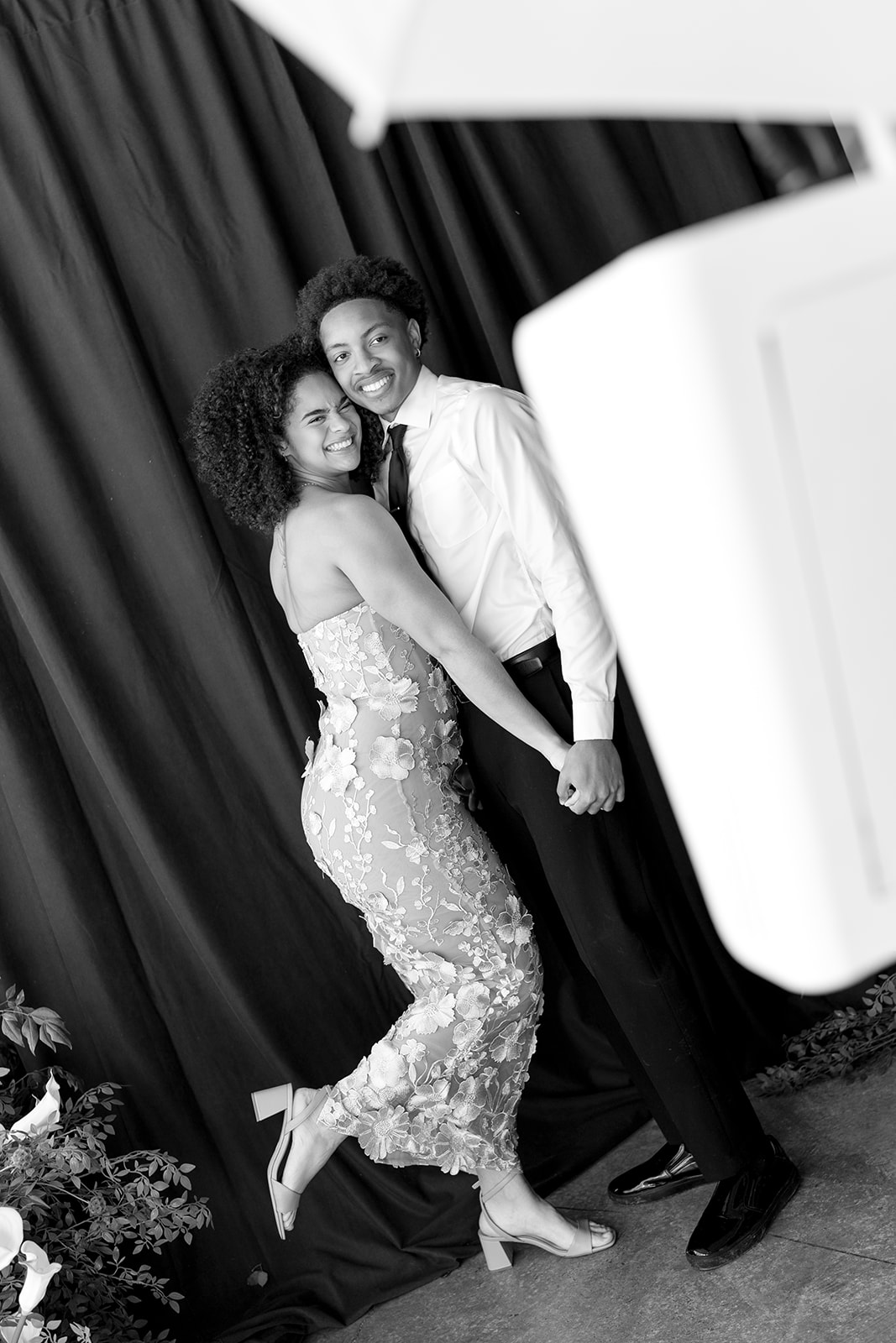 Black-and-white shot of a couple posing at a dark draped backdrop, the white booth blurred in the foreground